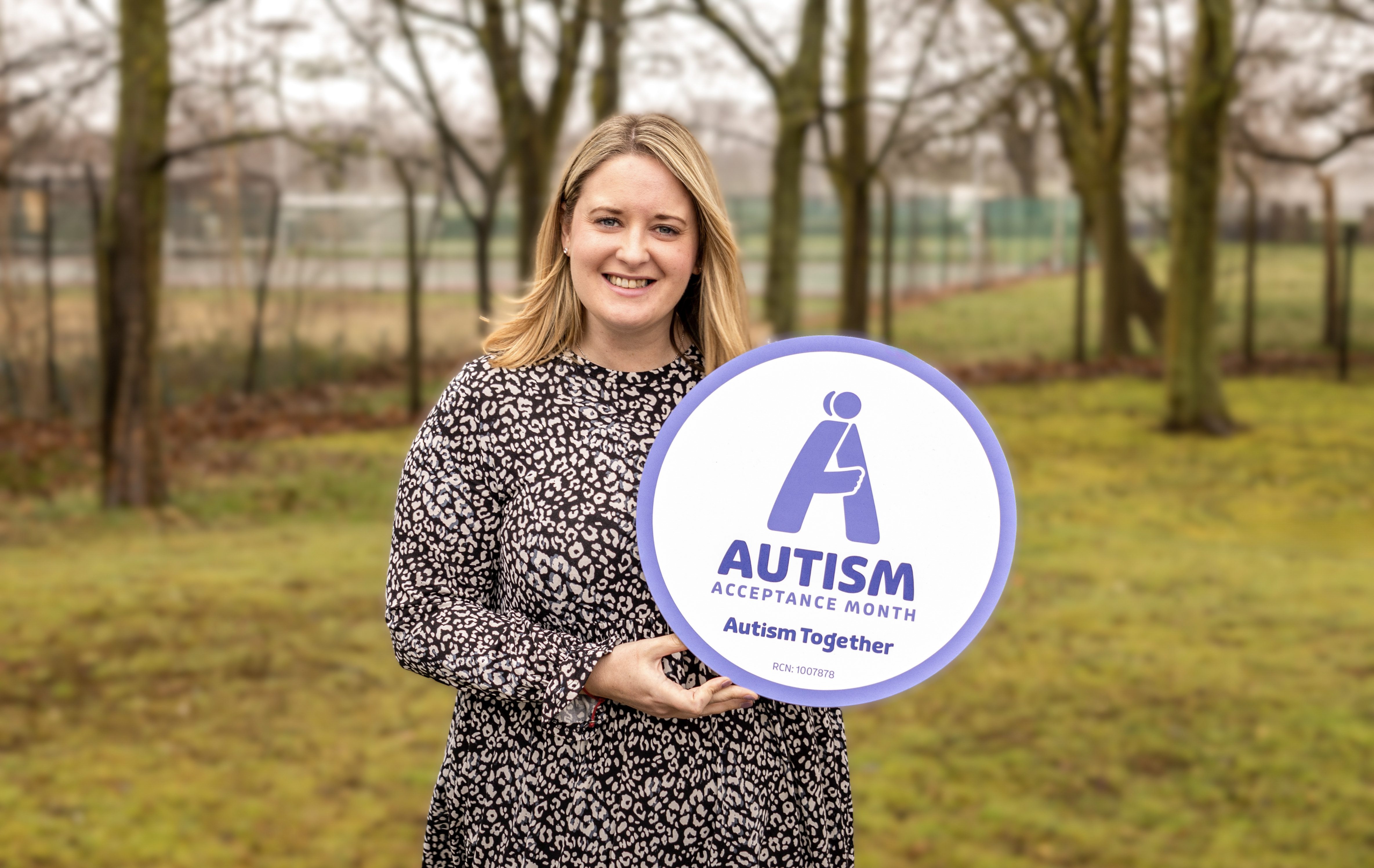 Woman with brown hair and a long frock in a grassy area holding a purple Autism Acceptance Month sign