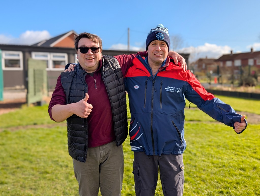 Two men standing in a green space with one-storey buildings behind. The man on the right is wearing Special Olympics Winter Games jacket and hat