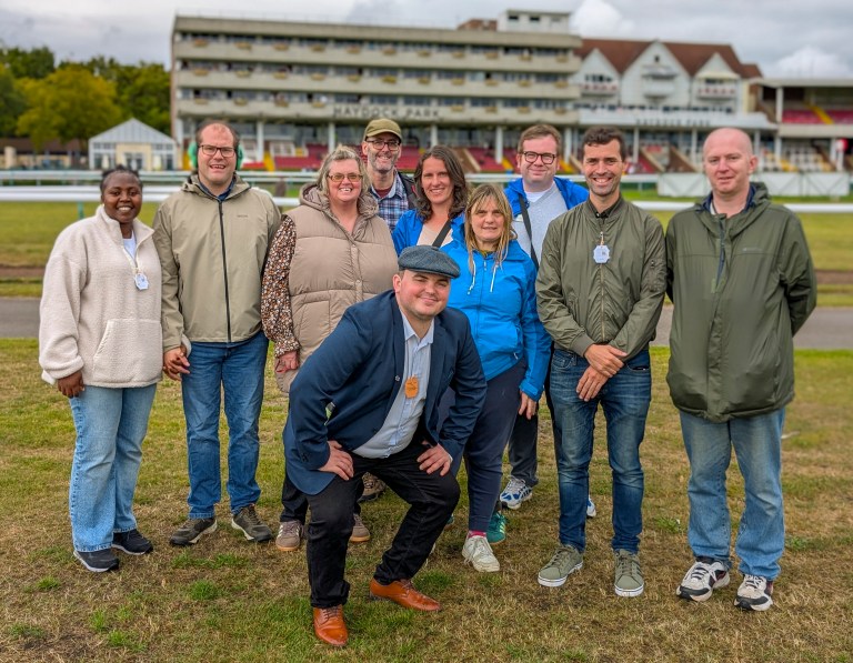 A group of people standing on a racecourse, with stands behind them on a grey day