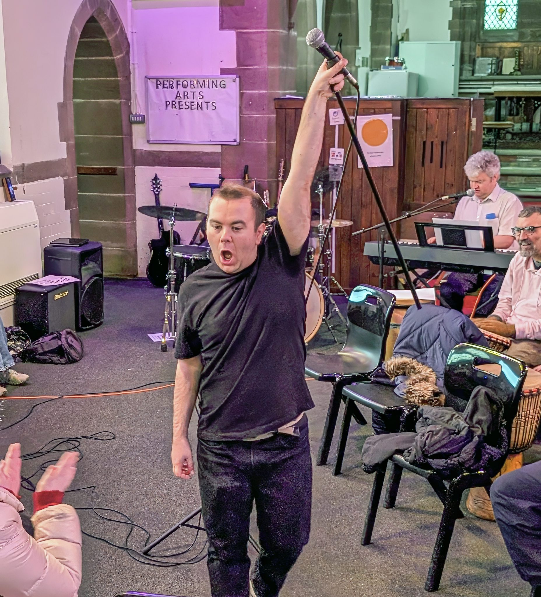 Man in a t-shirt and jeans holding his microphone and stand above his head as he performs with a band in an old church