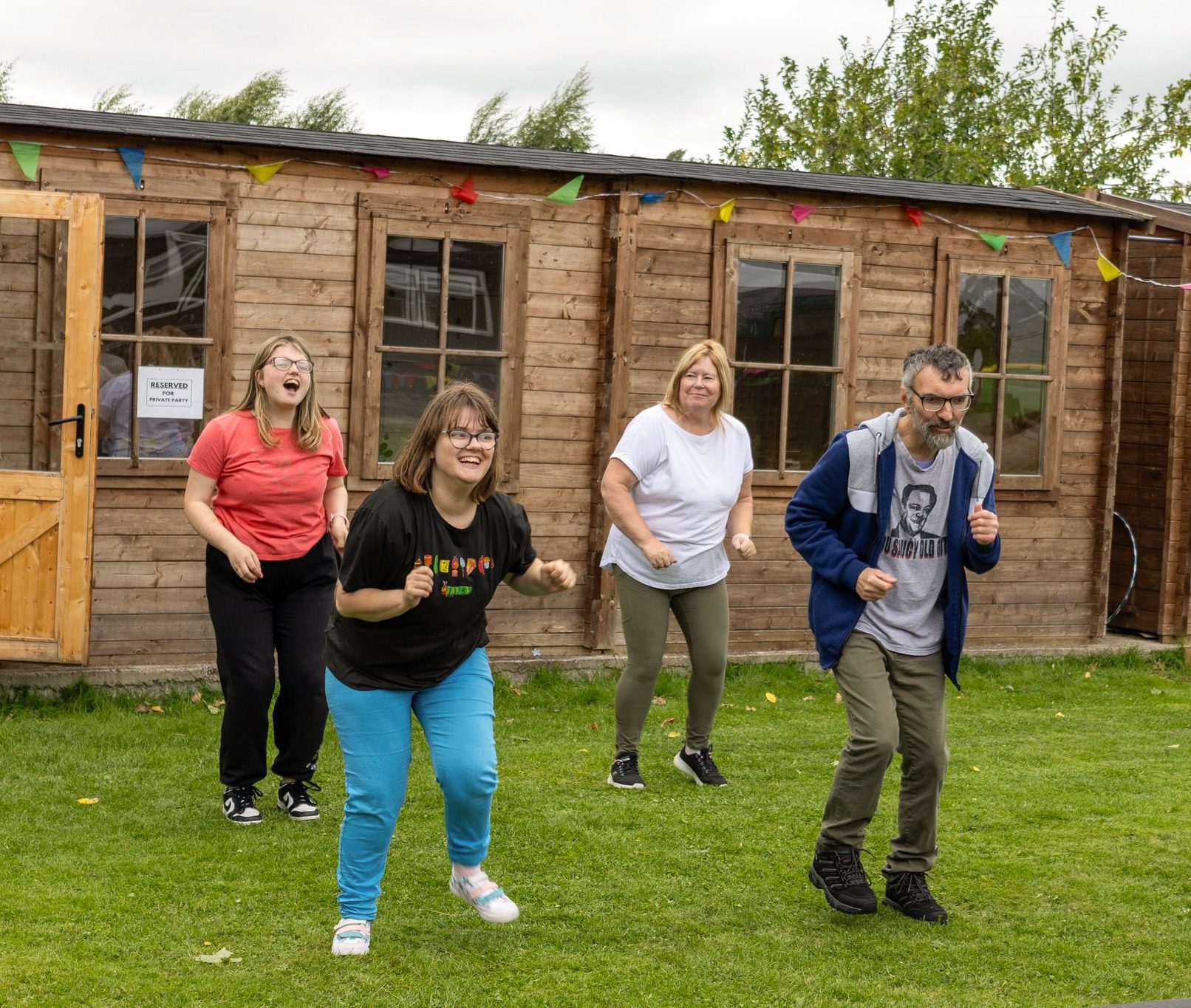 Dour people dancing together outside on the grass with wooden huts behind