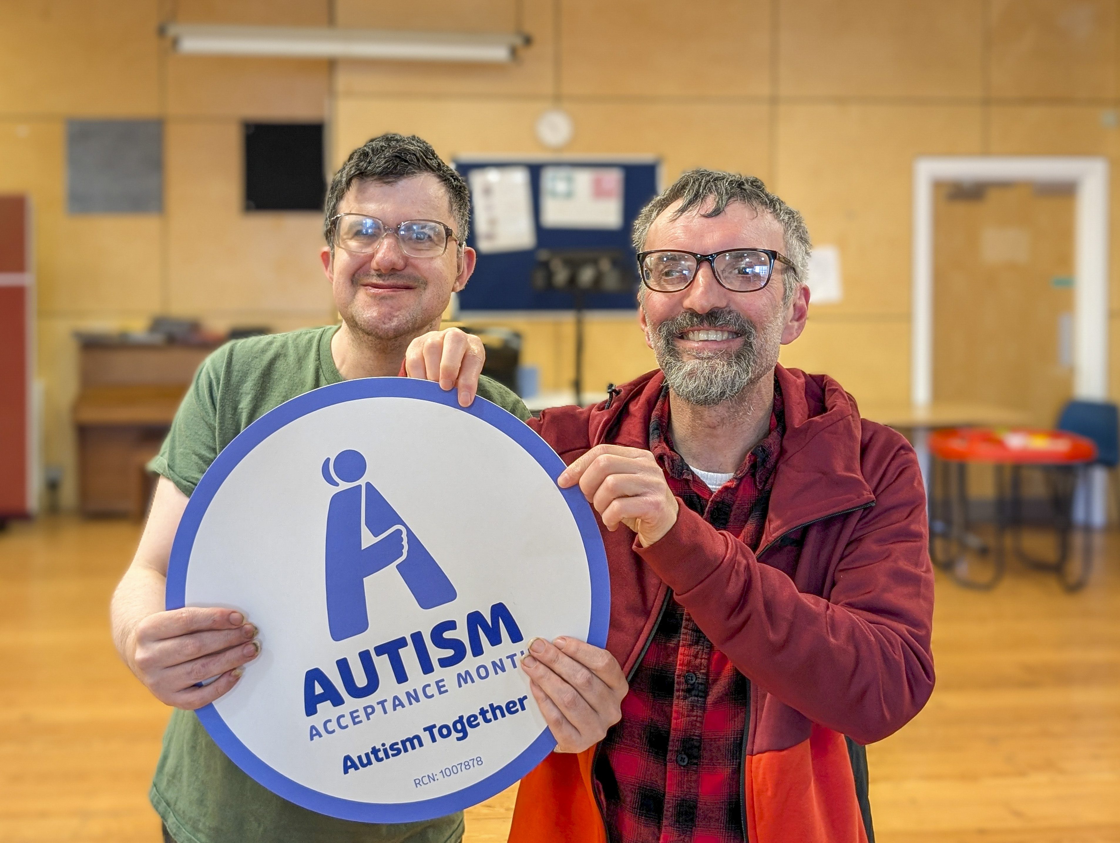 Two men; both with glasses, one with a beard, hold up an Autism Acceptance Month sign