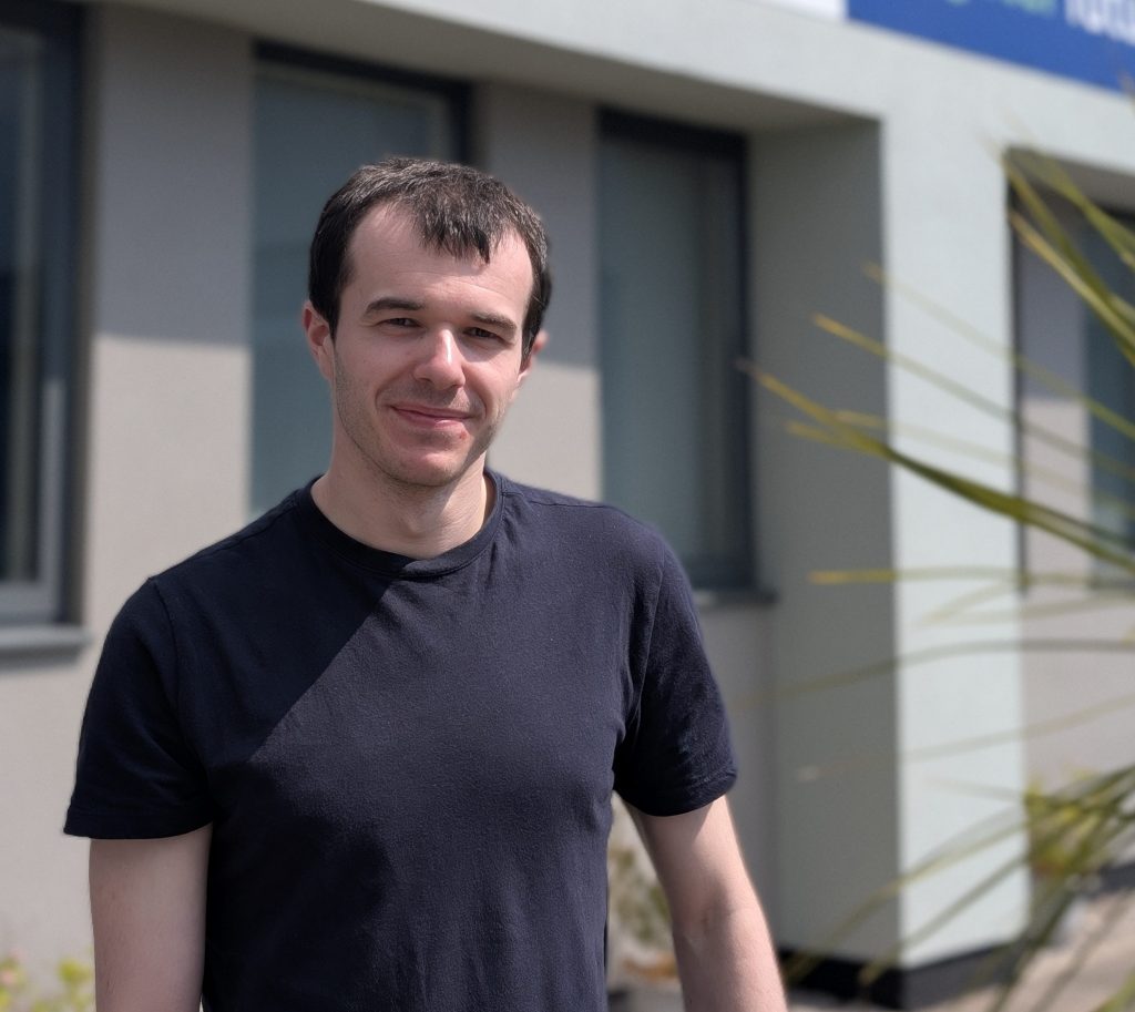 A young man in a black t-shirt stands outside a building with the logo for charity Autism Together above his head