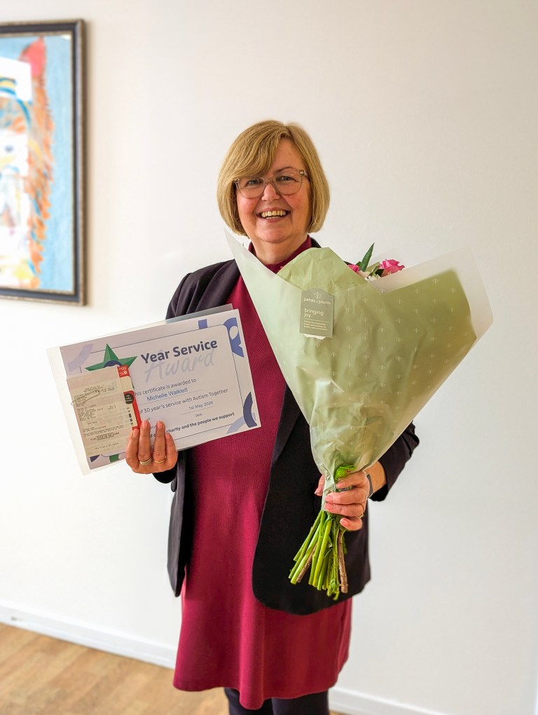 Smiling lady with a blonde bob and glasses in a corridor, holding flowers and a certificate of 30 years' service