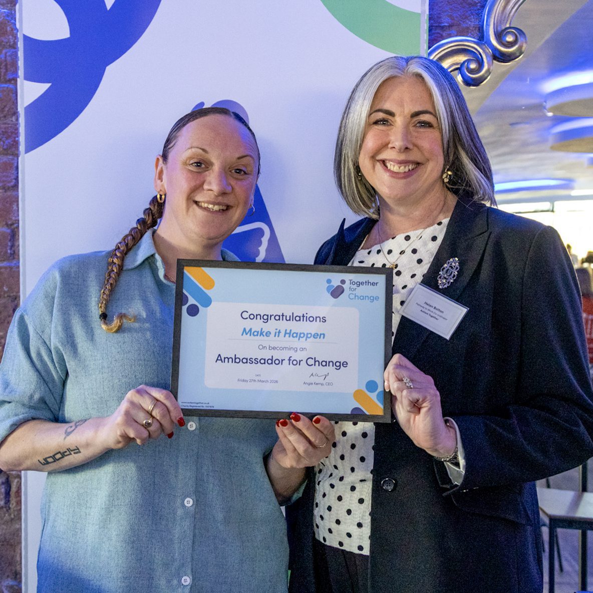 Two women in an event, standing before an Autism Together banner holding a certificate saying Make it Happen is an Ambassador for Change