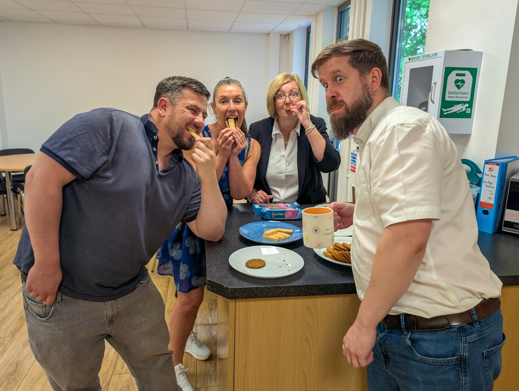 Four people, two men and two women, each tasting biscuits and taking on funny poses with biscuits in their mouths, standing around a worksurface with different biscuits on plates.