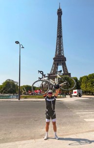 Man standing in the street in Paris. He is holding a bicycle above his head in jubilation and the Eiffel Tower is in the background