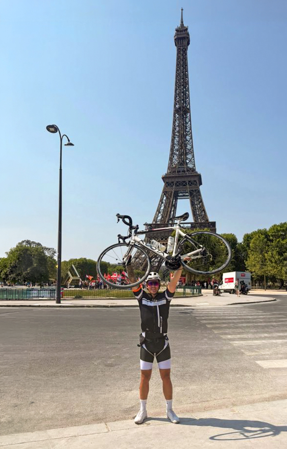 Man standing in the street in Paris. He is holding a bicycle above his head in jubilation and the Eiffel Tower is in the background